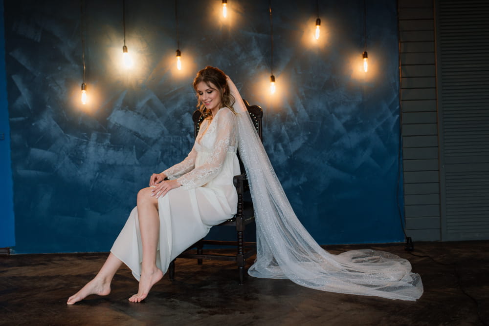 Woman in a white lace dress and sparkling veil sitting for a photoshoot against a blue wall with hanging Edison bulbs