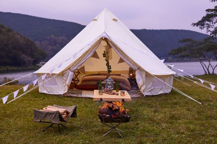 A spacious white canvas bell tent is pitched on a grassy field near a calm lake at twilight, offering a luxurious outdoor glamping experience with an inviting campfire and cozy interior visible.